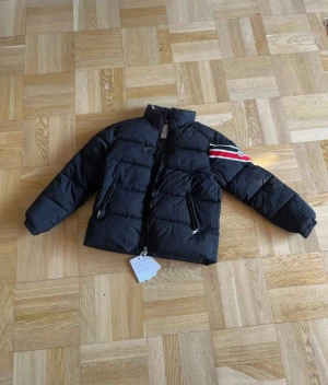 Svart Moncler pufferjacka med ränder - Svart pufferjacka från Moncler med coola röda och vita ränder på ena ärmen och Moncler-logga. Jackan har hög krage, två fickor med dragkedja och en snygg, quiltad design. Perfekt för dig som vill ha en varm och stilren jacka med streetkänsla.