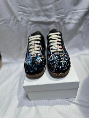 Maison Margielas - Maison Margiela sneakers i svart skinn med unikt färgstänksmönster i blått, vitt, rött och gult. Klassisk låg modell med rund tå, vita snören och gummisula i beige. Perfekt för dig som vill sticka ut med en konstnärlig vibe och exklusiv känsla. Box hänger med vid frågor är det att bara skriva!