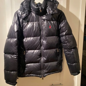 Svart pufferjacka från Polo Ralph Lauren - Svart pufferjacka från Polo Ralph Lauren med glansig finish och röd logga på bröstet.  Säljer pga för lite för mig, sitter mer som en small
