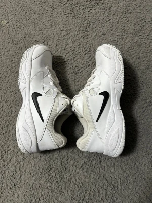 Nike Court Lite 2 vita sneakers - Nike Court Lite 2 sneakers i helvitt med svart swoosh på sidan. Skorna har en chunky siluett, snörning och ventilerande meshdetaljer. Yttermaterialet är en mix av skinn och syntet, med perforeringar för extra andningsförmåga. Perfekt för dig som gillar sportig och clean stil.