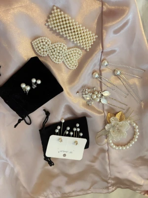 Pärlsmyckes-set med håraccessoarer - Säljer ett elegant set med pärlsmycken och håraccessoarer. Innehåller två stora hårspännen med pärlor, ett pärlarmband med vit blomma och guldfärgade blad, flera hårnålar med pärlor och silverdetaljer samt örhängen med pärlor och strass. Perfekt för att lyfta din look med glittriga detaljer.