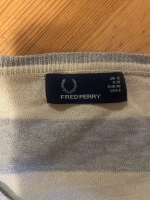 Randig v-ringad tröja Fred Perry - Snygg v-ringad tröja från Fred Perry i beige och grått med breda ränder. Tröjan har lång ärm och är stickad i mjukt material, perfekt för dig som gillar klassisk stil med en twist. Passformen är normal och den har diskret logga på bröstet.