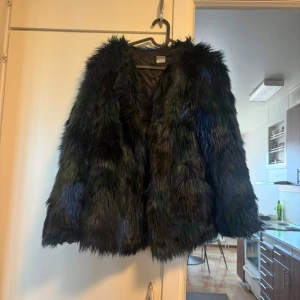 Svart pälsjacka med gröna inslag - Säljer en fluffig och cool pälsjacka i svart med mörkgröna och blå toner. Jackan har lång ärm och en oversized passform som ger en riktigt statement-look. Perfekt för dig som vill sticka ut och hålla dig varm under höst och vinter.