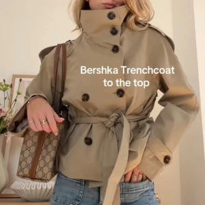Bershka trenchcoat - Snygg beige trenchcoat från Bershka med högt krage, knappar framtill och bälte i midjan. Jackan har en kortare, croppad passform och långa ärmar. Helt slutsåld online, kommer inte sälja den för mindre än 900kr. Helt ny och endast provad!