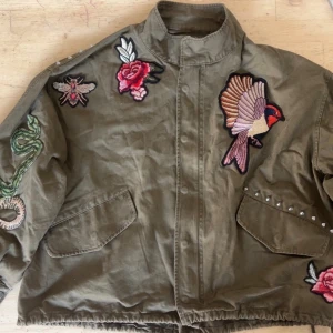 Grön jacka med broderier - Säljer en grön jacka med coola broderade detaljer som fågel, rosor, bi och orm. Jackan är i storlek xs men passar lätt vem som i S och skulle nog funka bra för M med. Jackan har en dragkedja, tryckknappar och nitar på axeln och vid fickan 