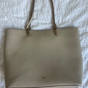 Beige axelväska i skinn - Stilren beige axelväska med guldfärgade kedjedetaljer och långa handtag. Väskan är tillverkad i slätt fakeskinn och har en minimalistisk design med diskret logga framtill. Perfekt för dig som gillar klassiska och enkla accessoarer. Väskan har dock defekter på banden, slitningar på kedjan och på loggan fram till.