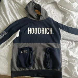Blå och grå HOODRICH hoodie med fickor - Säljer en blå och grå hoodie från HOODRICH med stor logga på bröstet och ärmen. Tröjan har huva, två stora fickor med dragkedja framtill och coola kontrastpaneler. Perfekt för dig som gillar streetwear och vill sticka ut med en unik look. Den har inga snören vid luvan 