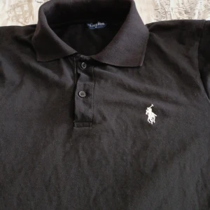 Svart pikétröja från US Polo Assn - Svart pikétröja från US Polo Assn med klassisk krage och knäppning framtill. Tröjan har korta ärmar och är prydd med den ikoniska broderade loggan i vitt på bröstet. Perfekt för en clean och stilren look.