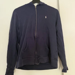 Mörkblå hoodie från Ralph Lauren - Mörkblå hoodie från Ralph Lauren med dragkedja framtill och klassisk huva. Tröjan har långa ärmar, känguruficka och den ikoniska lilla loggan broderad på bröstet. Perfekt för chill dagar eller när du vill ha en enkel och stilren look.