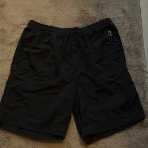 Svarta badbyxor från Parajumpers S - Svarta badbyxor från Parajumpers med elastisk midja och snörning. Klassisk design med diskret logga och metallring på sidan. Lätta och bekväma, perfekta för varma dagar. Tillverkade i syntetmaterial med meshfoder för extra komfort. Nypris 1700 kr 