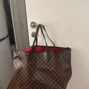 Neverfull väska - 31cm höjd, 54cm bredd! Lo*** V****** Neverfull i Damier-mönster med bruna och mörkbruna rutor. Väskan har svarta skinnhandtag som är lite slitna men inte så synligt, rödrandigt innerfoder och medföljande clutch. Perfekt storlek för allt du behöver ha med dig. 