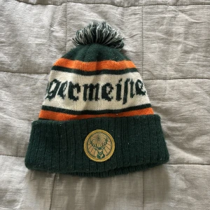 Jägermeister mössa med tofs - En knappt använd Snygg stickad mössa från Jägermeister i mörkgrönt, orange och vitt med bred uppvikt kant och broderad logga framtill. Mössan har en stor tofs på toppen och coolt randigt mönster med Jägermeister-tryck runt om. Perfekt för dig som gillar streetwear och vill sticka ut.