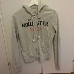 Vit kofta från  Hollister - Vit kofta från Hollister med dragkedja framtill, broderad logga och text i svart och rosa på bröstet. Tröjan har huva med snörning, två fickor fram och ribbade muddar. Perfekt för chill dagar och enkel att matcha med jeans.