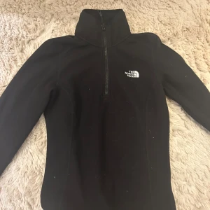 Svart fleece från The North Face - Svart fleece-tröja från The North Face med halv dragkedja och hög krage. Tröjan har lång ärm och en broderad logga på bröstet. Perfekt för kyliga dagar och outdoor vibes.