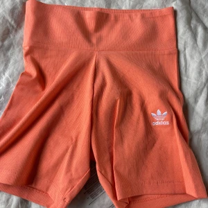 Coral ribbade shorts från Adidas - Snygga orange ribbade shorts från Adidas med hög midja och vit broderad logga framtill. Perfekta för träning eller chill dagar. Stretchigt material som sitter skönt och följer kroppen. Fräsch färg som sticker ut och ger en sportig vibe.