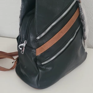  Helt ny Svart ryggsäck från Amalia med bruna detaljer -  Helt ny Snygg svart ryggsäck från Amalia med bruna axelremmar och detaljer. Väskan har flera dragkedjefack framtill och på sidorna, vilket gör den både praktisk och stilren. Tillverkad i syntetmaterial med silverfärgade dragkedjor och justerbara remmar. 33x28x13 