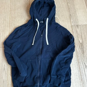 Intimissimi (kille) mörkblå hoodie - Mörkblå hoodie med vit snörning i huvan och hel dragkedja framtill. Tröjan har långa ärmar, ribbade muddar och två fickor framtill. Perfekt för en avslappnad stil och enkel att matcha med jeans eller joggers.