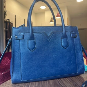 Blå handväska från Valentino i skinn - Snygg blå handväska från Valentino i slätt skinn med stilren design och V-detalj framtill. Väskan har både handtag och axelrem, samt dragkedja upptill och flera fack inuti. Perfekt för dig som vill ha en klassisk och trendig accessoar. Kan ej garantera att den är äkta men tror det, därav de billiga priset