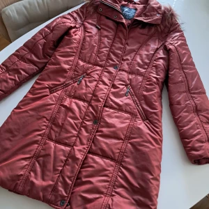 Rostfärgad lång dunjacka från Luhta - Lång, quiltad dunjacka från Luhta i en snygg rostig röd färg. Jackan har huva med päls, dragkedja och knappar framtill samt två dragkedjefickor. Perfekt för kalla dagar med sin varma och mjuka känsla.