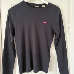 Svart långärmad tröja från Levi's - En stilren svart långärmad tröja från Levi's med klassisk rund halsringning och den ikoniska röda Levi's-loggan på bröstet. Tillverkad i mjuk bomull som är skön mot huden. Perfekt för dig som gillar enkel och tidlös stil.