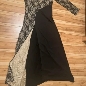 Svart och beige långklänning med spets - Unik långklänning i svart och beige med asymmetrisk design. Ena sidan har ett snyggt spetsmönster över beige tyg, medan andra sidan är helsvart. Långa ärmar och elegant slits framtill. Perfekt för dig som vill sticka ut med en cool och sofistikerad look.