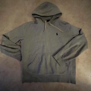 Grå hoodie från Ralph Lauren - Säljer en klassisk grå hoodie från Ralph Lauren med broderad logga på bröstet. Tröjan har huva med snörning, magficka och långa ärmar. Perfekt för dig som gillar en enkel och stilren look.