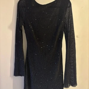 Glittrig kortklänning med öppen rygg - Supersnygg svart kortklänning med glittriga detaljer och lång ärm. Klänningen har en djup öppen rygg med knytband och sitter figurnära. Använd en gång