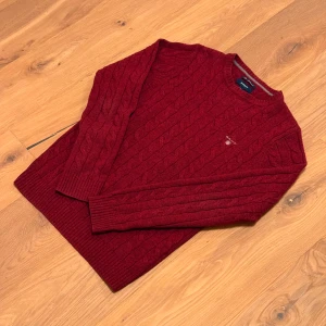 Vinröd kabelstickad tröja från GANT - Vinröd kabelstickad tröja från GANT i mjuk lammull. Klassisk rund halsringning, ribbade muddar och broderad GANT-logga på bröstet.