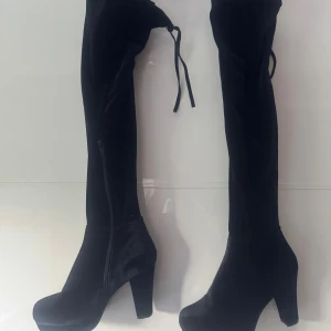 Svarta overknee stövlar med klack - Säljer ett par svarta overknee stövlar med hög klack och rund tå. Skorna är i mjuk mocka-liknande syntet, har dragkedja på insidan och knytning upptill för perfekt passform. Perfekta för dig som vill ha en edgy och trendig look. Använd en gång.