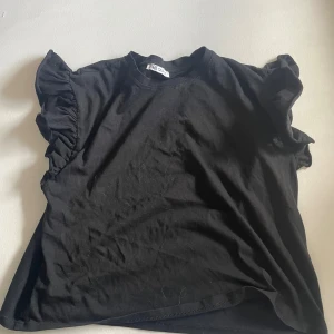 Svart volangblus från Zara - Svart blus från Zara med korta ärmar och snygga volangdetaljer på axlarna. Rund halsringning och mjukt bomullsmaterial som känns skönt mot huden. Perfekt för dig som gillar en enkel men ändå trendig look.