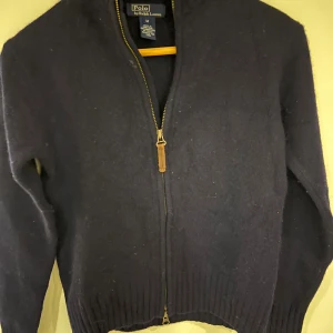 Mörkblå kofta från Polo Ralph Lauren - Mörkblå kofta från Polo Ralph Lauren i 100% lammull. Tröjan har hel dragkedja framtill och ribbade muddar vid ärmslut och nederkant. Perfekt för dig som gillar klassisk stil med en modern touch. OBS passar s/xs det står M i amerikansk ungdoms stl