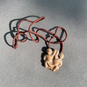 Handgjort halsband med lerhänge - Unikt halsband med ett handformat hänge i lera som föreställer två figurer. Hänget har en naturlig beige färg och hänger i ett brunt snöre. Perfekt för dig som gillar personliga och kreativa accessoarer.