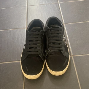 Svarta Saint Laurent sneakers - Svarta sneakers från Saint Laurent med vit textlogga på sidan. Skorna är i ett 7/10 skick då de är tråd(se på bild ) på skorna vilket gör att de blir ett litet hål under tråden, ingenting som syns och går att sy igen. Dustbag följer med men tyvärr ingen box. Pris går att diskuteras vid snabb affär, tveka inte på att höra av er om tex fler bilder eller liknande. Fraktar väldigt fort. Strl 41