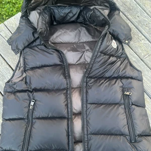 Svart dunväst från Moncler med huva - Säljer en svart dunväst från Moncler med avtagbar huva och två dragkedjefickor framtill. Västen har quiltad design och klassisk Moncler-logga på bröstet. Perfekt för lager-på-lager och håller dig varm under kyliga dagar. Storlek 1