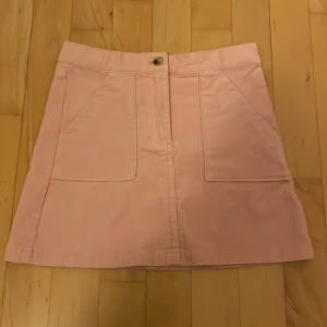 Ljusrosa manchesterkjol från H&M - Supersöt ljusrosa kortkjol i manchester från H&M. Kjolen har två stora fickor framtill, knapp och dragkedja i midjan. Perfekt för dig som gillar en enkel men trendig look med retrovibbar.