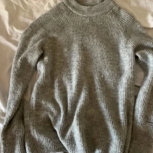 Grå stickad tröja med hög krage - Mysig grå stickad tröja med lång ärm. Tröjan har en ribbad struktur och är perfekt för kyliga dagar. Enkel och stilren design som passar till det mesta.