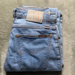 Ljusblå jeans från Nudie Jeans, 29/30 - Säljer ett par ljusblå jeans från Nudie Jeans i klassisk femficksmodell. Jeansen har orange kontrastsömmar, patch i mockaimitation bak och snyggt broderad bakficka. Perfekta för dig som gillar en clean och tidlös look.