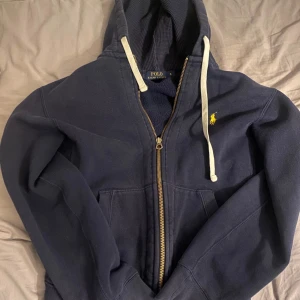 Marinblå hoodie från Polo Ralph Lauren - Säljer en marinblå hoodie från Polo Ralph Lauren med dragkedja och huva. Tröjan har vit snörning i huvan, gult broderat logotyp på bröstet och är tillverkad i mjuk bomull.