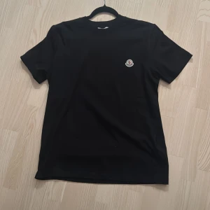 Svart t-shirt från Moncler - Svart t-shirt från Moncler med klassisk passform och rund hals. T-shirten har korta ärmar och en liten Moncler-logga broderad på bröstet. Perfekt för en clean och stilren look.