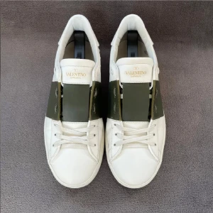 Valentino Garavani Open sneakers vit/grön - Snygga Valentino Garavani Open sneakers i vitt skinn med bred grön rem över mitten. Klassisk rund tå, platt sula och diskret logga på plösen. Skorna har snörning och coola nitar baktill på sulan. Perfekta för dig som gillar exklusiva och stilrena sneakers.