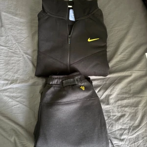 Nike NOCTA svart tracksuit  - Svart hoodie fràn Nike NOCTA med gul logga och detaljer. Tröjan har huva, dragkedja framtill som ger en modern look. Materialet är mjukt och skönt, perfekt för chill eller streetwear. NOCTA-tryck pả ärmen och bröstet i gult Samt byxorna 