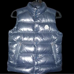 Blå dunväst från Moncler - Säljer en glansig blå dunväst från Moncler med hög krage och två dragkedjefickor framtill. Västen har quiltad design och klassisk Moncler-logga på bröstet. Perfekt för lager-på-lager och snygg till streetwear. Den är storlek 152 cm men passar även större