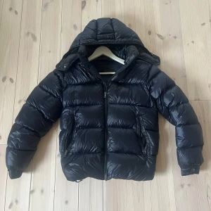 Svart pufferjacka från Massimo Dutti - Snygg svart pufferjacka från Massimo Dutti med huva och dragkedja framtill. Jackan är vadderad och har två fickor med dragkedja på sidorna. Perfekt för kalla dagar när du vill hålla värmen och ändå se stilren ut.