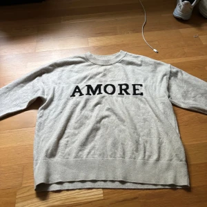 Beige stickad tröja med AMORE-tryck - Beige stickad tröja med svart texten 'AMORE' framtill. Tröjan har rund halsringning och långa ärmar. Perfekt för dig som gillar enkel men snygg stil med en statement-detalj.