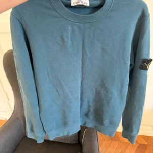 Blå sweatshirt från Stone Island - Säljer en blå sweatshirt från Stone Island med klassisk rund hals och ribbade muddar. Tröjan har den ikoniska loggan på vänster ärm och är tillverkad i mjuk bomull. Perfekt för dig som gillar streetwear och snygga detaljer. Tyvärr är lapparna på insidan bortklippta av förra ägaren som tyckte det kliade.