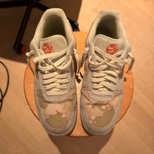 Nike Air Force 1 Camo Beige Sneakers - Unika Nike Air Force 1 sneakers i beige och ljusgrått med camouflagemönster. Skorna har klassisk snörning, vit swoosh och orange Nike-logga på plösen. Materialmix av mocka och textil ger en cool och modern look.