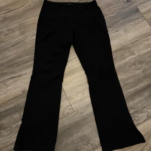 Svarta bootcut kostymbyxor med slits - Snygga svarta kostymbyxor med bootcut-snitt, slits längst ner och klassisk passform. Byxorna har hällor för bälte och stängs med dragkedja och knapp. Perfekta för en stilren look och passar till många olika outfits.