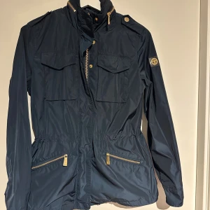 Mörkblå parkajacka med guldiga detaljer - Parkajacka i mörkblått från Morris med guldiga dragkedjor och knappar. Jackan har fyra fickor framtill, hög krage och justerbar midja. Materialet är lätt och vattenavvisande polyester, perfekt för vår och höst. Klassisk badge på ärmen och snygga detaljer.