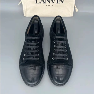 Svarta sneakers från Lanvin i mocka - Snygga svarta sneakers från Lanvin med ovandel i mjuk mocka och tåparti i präglat skinn med krokodilmönster. Skorna har rund tå, svarta snören och platt sula. Perfekta för dig som gillar stilrena och lyxiga detaljer.