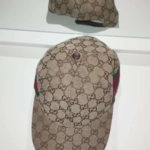 Gucci keps med GG-mönster - Snygg keps från Gucci i beige med klassiskt GG-monogram över hela kepsen. Har gröna och röda ränder på sidan och justerbar rem baktill. Tillverkad i bomullsmix med insida i vitt tyg. Perfekt för dig som vill sticka ut med en lyxig accessoar.